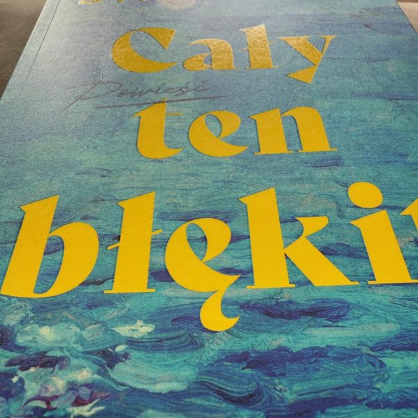 Widok z perspektywy żabiej na dolną część okładki książki "Cały ten błękit". Dominują jaskrawożółte litery tytułu na tle turkusowo-niebieskiej grafiki przypominającej fale.
