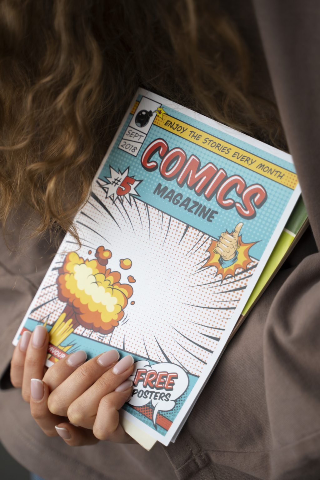 Zbliżenie na dłonie osoby trzymającej kolorowy zeszyt komiksowy o tytule "COMICS MAGAZINE". Na okładce widoczna jest grafika przedstawiająca wybuch w stylu pop-art oraz kciuk uniesiony w górę.