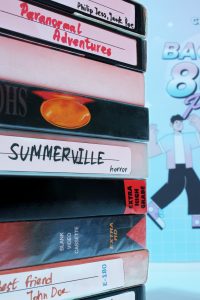 Stos kaset wideo VHS ułożonych jedna na drugiej. Na etykietach widoczne są odręczne napisy takie jak "Paranormal Adventures", "Summerville horror" oraz "Best friend John Doe". W tle widać fragment plakatu w stylistyce lat 80.