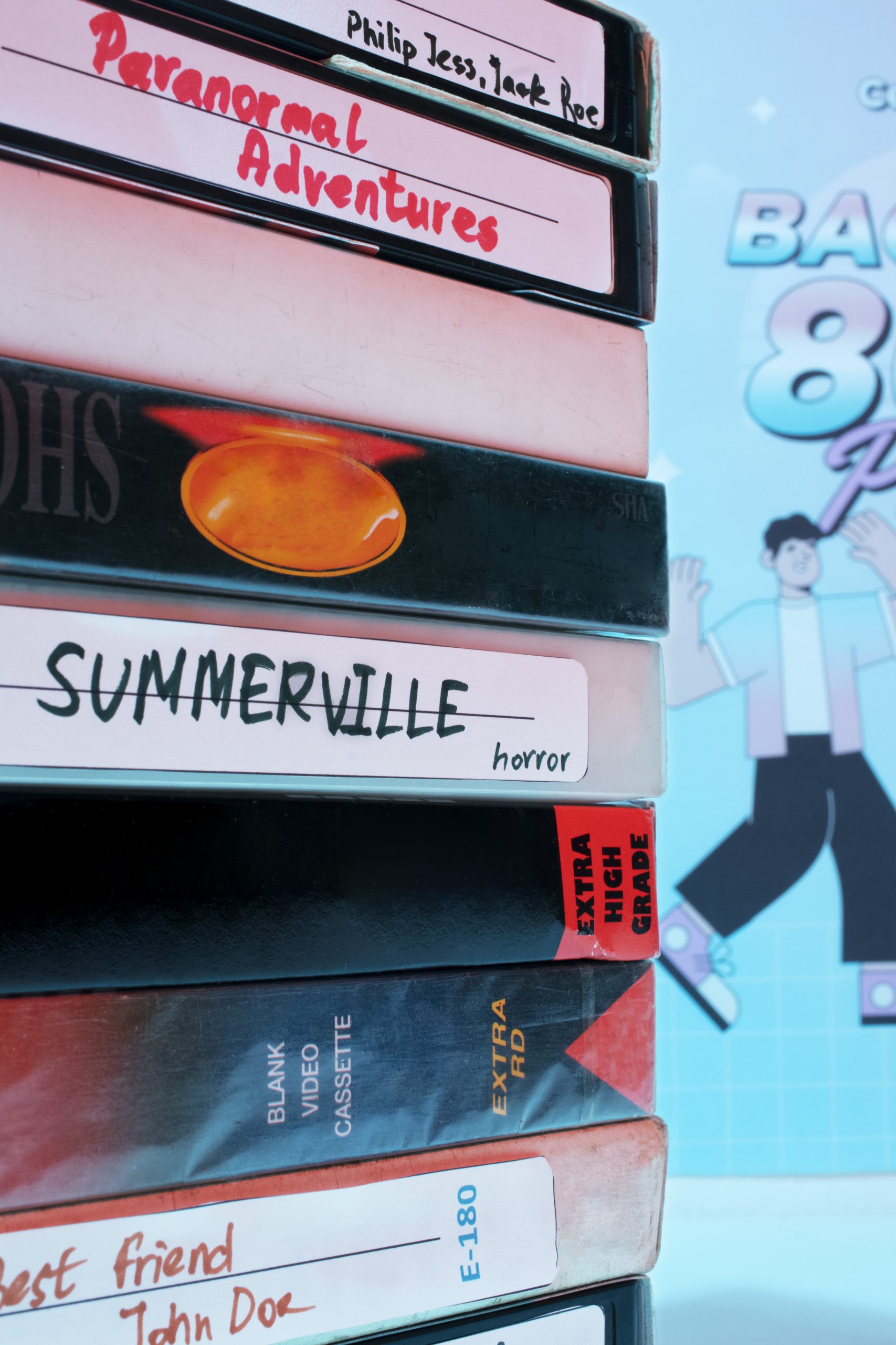 Stos kaset wideo VHS ułożonych jedna na drugiej. Na etykietach widoczne są odręczne napisy takie jak "Paranormal Adventures", "Summerville horror" oraz "Best friend John Doe". W tle widać fragment plakatu w stylistyce lat 80.