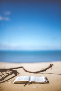 Otwarta książka leżąca na piaszczystej plaży. W tle widać rozmyty błękit oceanu i jasne niebo, a obok książki leży kawałek suchego drewna wyrzuconego przez morze.